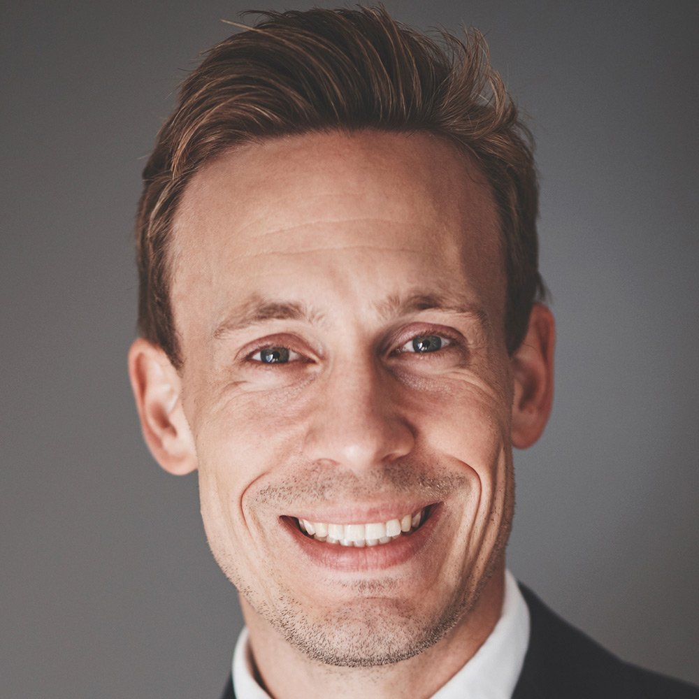 A man with light brown hair, blue eyes, and a wide smile is wearing a dark suit and white shirt, facing the camera against a plain gray background.